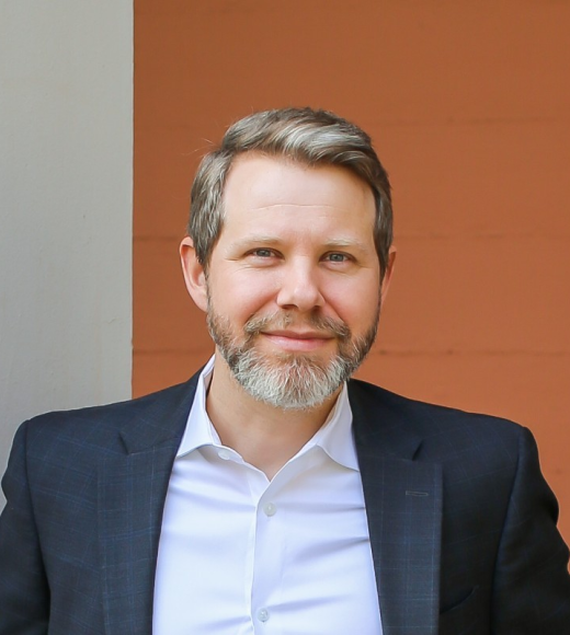 White man with greying hair and beard smiles at camera, wearing a white buttoned-down shirt and dark sports coat.light